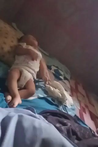 The video shows a close-up shot of a baby lying on a bed. The baby is wearing a white onesie with a cartoon print and a diaper. The baby's head is resting on a pillow, and their eyes are open, looking upwards. The baby's left arm is resting on their chest, while their right arm is extended to the side. The bed is covered with blankets and sheets in various colors and patterns, including blue, white, and patterned fabric. A pile of white cloth is visible next to the baby. The background is dark and appears to be the inside of a room, possibly a bedroom. The lighting is dim, casting shadows on the scene. The camera angle is slightly above and to the side of the baby, focusing on the baby's face and upper body.