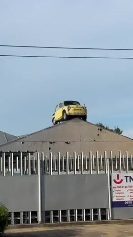 The video shows a yellow, vintage car parked precariously on the peak of a corrugated metal roof. The car is angled, with its front facing slightly left. The roof itself appears weathered and has several visible holes. Below the roof, a silver metal fence with vertical slats runs across the bottom of the frame. To the right of the fence, a sign with red lettering and a red symbol is partially visible. The sky is clear with a light blue hue. The car's license plate is visible, reading "KLE-998 D". The overall impression is that of an artistic installation or an unusual advertisement.