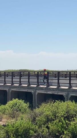 The video shows a man walking across a bridge in a natural environment. The man is wearing a red hat and a multi-colored shirt. The bridge is constructed of concrete and has metal railings. There is green vegetation surrounding the bridge, and a small river flows underneath it. The sky is blue with a few white clouds. The overall scene appears to be a sunny day in a rural or natural setting.