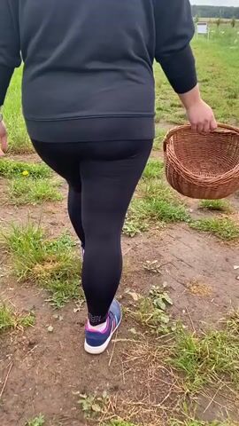 A person walks away from the camera, carrying a wicker basket in their right hand. They are wearing black leggings with a logo on the left calf, a black sweatshirt, and athletic shoes. The person is walking on a dirt path through a grassy field, with more greenery and a hint of trees in the background. The sky is overcast.