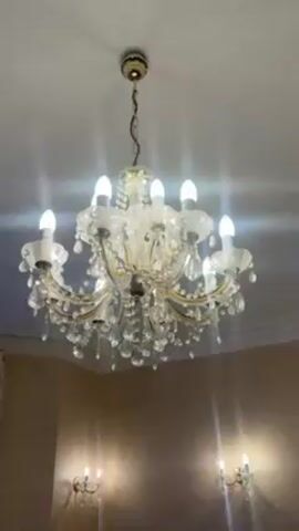 The camera pans slowly around a dining room, moving from a chandelier with crystal pendants, to a window with elaborate floral drapery, and then to a fireplace with a decorative mirror above it. The room has light yellow textured wallpaper. The window looks out on a view of bare trees and rooftops. The room has several decorative wall sconces with candle-like bulbs. Someone speaks and comments on the prettiness of the room.