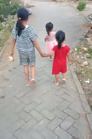 A brief video shows three young girls walking on a paved path. The tallest girl, wearing a patterned onesie, walks in the foreground, holding the hands of the two younger girls. The middle girl wears a pink dress, and the youngest girl wears a red dress. They are walking away from the camera, down the path. The environment appears to be a rural or suburban area, with some vegetation and buildings visible in the background. The lighting suggests it is daytime.