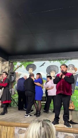 The video shows a group of people dancing on a stage. There are about 10 people on stage, with a man in a red vest singing into a microphone in the foreground. Behind him, couples are dancing in a circle. To his left, a couple dressed in traditional clothing is dancing. The backdrop of the stage is a colorful mural depicting people and trees. A guitar leans against the back wall as well. The stage is lit and appears to be in a restaurant or club.