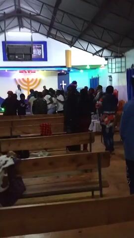 The video shows a church service in progress. People are standing and moving in the area in front of the altar. The altar is decorated with a menorah, crosses, and colored lights. There are wooden pews in the foreground, with some items left on them. The building has a high roof.