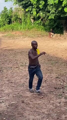 The video features two men dancing outdoors on an unpaved, dirt ground. The setting is rural, with lush green trees and foliage in the background.

The main subject is a shirtless man with a bald head, wearing blue jeans and sandals, with a yellow shirt draped over his left shoulder. He is actively dancing, moving his arms and legs in rhythm with the music. Another man, wearing a light grey polo shirt and blue jeans, approaches him and also begins to dance, facing the shirtless man. Both men appear to be enjoying themselves, with focused and engaged expressions.

In the background, a third person is seated on a chair under the shade of a tree. The ground is dry and uneven. The scene is well-lit, suggesting it is daytime. No visible text is present. The activity seems to be a social gathering or a performance, characterized by joyful dancing.