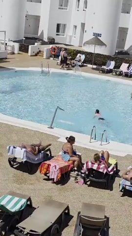The video shows a sunny outdoor scene at a swimming pool. Several people are visible. In the foreground, four lounge chairs are occupied by sunbathers. One person lies on their stomach, another sits up, and two lie on their backs. A woman in a blue swimsuit is sitting in one chair, and a blonde woman is reclining in another. Colorful towels are draped over the chairs. In the pool, a man is swimming, and a group of three people, two adults and a child, are standing in the shallow end. Further back, near the edge of the pool, several lounge chairs are set up, some occupied by people. A tall white building with multiple windows and balconies is visible in the background. The ground around the pool is covered in gravel and light-colored paving stones. A palm tree grows near the pool's edge. The water in the pool is clear and blue.