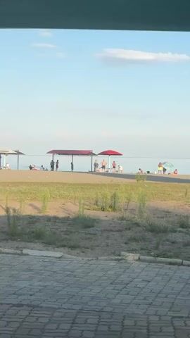 The video shows a beach scene with people scattered along the shore. Two canopies, one longer and one smaller, provide shade. Under the larger canopy, several people are gathered, some appearing to be adults and children interacting. One man is standing in front of the canopy, facing the camera. Another group of people are under a red umbrella further down the beach. There are also individuals sitting or standing near the water's edge. The environment appears to be a sunny day with a clear blue sky and a calm body of water in the background. The foreground consists of sand and sparse vegetation.