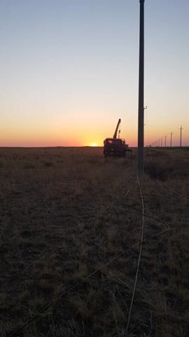The video shows a truck with a crane attached to it in a field at sunset. The sun is low on the horizon, casting a warm glow over the scene. The truck is parked next to a tall pole, and there are some wires running along the ground. The field is covered in dry grass, and there are more poles in the distance, forming a line across the landscape. The sky is a soft gradient of orange and purple.