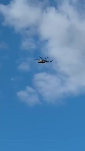 A helicopter flies across a blue sky with white clouds. The helicopter is dark and appears to be moving from left to right. The rotors of the helicopter are visible and spinning.