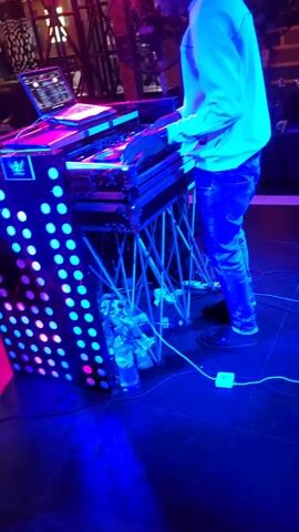 The video shows a DJ at work, operating a mixing console on a stage with changing colored lights. The DJ is a man wearing a long-sleeved white shirt, jeans, and sandals. He stands behind a DJ booth with a black front decorated with glowing white circles. A laptop is open on top of the booth, presumably connected to the console. The booth is supported by a framework of light-colored metal rods.  A bottle of water and some other items are visible beneath the booth. Other people are visible in the background, some of whom appear to be seated, and one other person walks by the DJ booth. The stage lights transition through various colors including blue, green, purple, and pink, creating a vibrant atmosphere. The floor of the stage is dark.