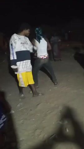 The video depicts an outdoor gathering at night. Several individuals are visible, some of whom are dancing to music. One person is wearing a white tank top and jeans, with a headscarf. Another person is wearing a white and blue top with yellow shorts. The background is dark, with some indication of uneven ground. There's a shadow of the camera-person visible in the foreground. The overall scene seems to be an informal social event.