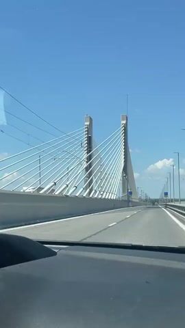 A car drives over a cable-stayed bridge on a sunny day. The bridge has two tall, concrete towers on either side of the road, from which numerous white cables extend down to the bridge deck, supporting it. The road is asphalt with white lane markings. There are also streetlights and some road signs visible along the bridge. The sky is clear blue with a few scattered white clouds. The camera is positioned inside the car, looking out through the windshield, capturing the perspective of the driver or a passenger.