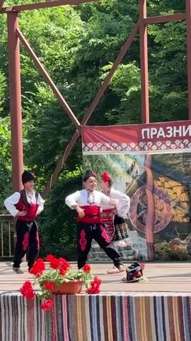 Three children in traditional Bulgarian folk costumes are performing a dance on an outdoor stage. The child on the left is wearing a black hat, white shirt, red vest, and black pants with red trim. The child in the middle is wearing a white blouse with embroidered details, a red and white patterned apron, and a flower in her hair. The child on the right is wearing a black hat, white shirt, red vest, and black pants with red trim. They are all smiling and appear to be enjoying themselves. The stage is decorated with a colorful striped fabric and potted red flowers. In the background, there is a banner with text in Cyrillic and a backdrop depicting a rustic scene with stone structures. Trees and greenery are visible behind the stage. The lighting suggests it is daytime, with sunlight filtering through the trees.