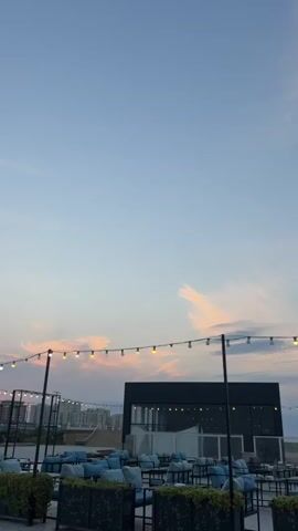 The video shows an outdoor seating area, possibly a restaurant or cafe, during what appears to be sunset. The sky is a pale blue with streaks of pink and orange clouds. String lights with round bulbs are hung across the scene. In the foreground, there are numerous tables and chairs with blue cushions, some covered by planters. Behind these, a dark, boxy building with lights visible inside is situated. In the middle ground and background, there are other structures, possibly apartment buildings, and further back, a body of water, likely the sea, is visible. Several people are present, mostly seen from behind or the side, sitting at tables or standing around. The atmosphere seems relaxed and casual.