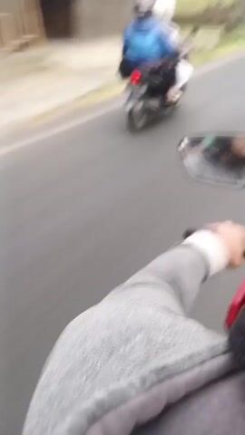 This is a first-person perspective video from a motorcycle on a paved road. The motorcycle is red and the rider is wearing a grey jacket. The rider's hand is visible on the handlebars. In front of the motorcycle, there are other vehicles and people. To the left, there is a scooter with two people on it, both wearing helmets and one wearing a blue jacket. Further ahead, there is a blue utility vehicle with several people standing in the back, some appearing to be wearing school uniforms and backpacks. There are also other motorcycles visible in the distance. The environment appears to be semi-urban or rural with trees and buildings along the side of the road. The sky is overcast. The video is shaky, indicating movement.