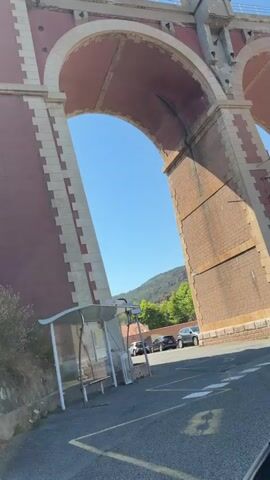 The video is shot from the perspective of someone riding in a vehicle, likely a car or motorcycle, as indicated by the slight shaking and the visible portion of a vehicle's interior in the bottom right corner. The vehicle is traveling along a paved road. To the left of the road, there is a vegetated embankment with a stone wall and a metal railing. Overhead, a large, multi-arched brick viaduct dominates the upper portion of the frame, with train tracks visible on top. The viaduct is constructed with red brick and has lighter colored stone accents at the edges of the arches and along the top. The arches are large and create shadows on the road and embankment. Beyond the viaduct, the sky is clear and blue, and there are some trees and hills visible in the distance. The sun is shining, casting shadows and illuminating the scene. There is a bus stop shelter with a bench visible on the right side of the road, and a car is parked near it. The video captures a dynamic perspective as the vehicle moves, showing the viaduct from different angles as it passes underneath.