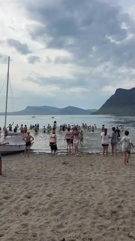 A group of people are gathered on a sandy beach and wading into the water. The weather appears overcast with dramatic clouds. Mountains are visible in the background across a body of water. Several individuals are already in the water, some swimming and others standing. A sailboat is docked on the left side of the frame. The people are casually dressed, many in swimwear. The overall impression is of a recreational gathering at a beach or lake.