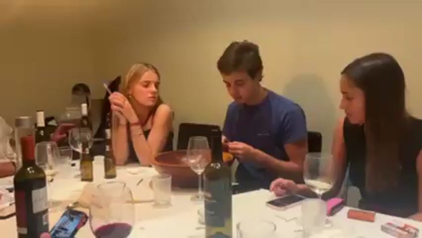 A group of approximately four people are seated around a table, possibly eating a meal. Two women and a man are prominently visible. The man, wearing a blue t-shirt, is speaking and gesturing with a small object in his hands. The women are listening to him. The table is cluttered with wine bottles, glasses, and other objects. The setting appears to be indoors, possibly a dining room or living space.