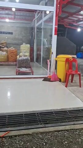 The video presents a stationary shot of the exterior of a small shop or mini-market at what appears to be dusk or night. The shop features large glass windows and doors framed in white, with internal red structural elements. Inside, multiple shelves and display racks are visible, stocked with various packaged goods, including snacks and other food items. On the left side of the interior, a tall, grey commercial refrigerator or display unit is situated. In the foreground, a metal grate covers a ground-level drain, with a rough, dark ground surface beyond it. The area directly outside the shop entrance has light-colored tiled flooring. To the far right, outside the shop, a yellow trash can and a red plastic chair are visible, along with a pink broom leaning against the wall. The shop's interior is brightly lit, illuminating the merchandise.