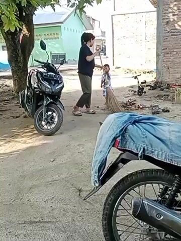 A woman is sweeping the ground with a broom near a black motorcycle parked under a tree. A young child is standing near the woman and holding the broom. The woman is wearing dark clothing and loose pants. The child is wearing a light-colored outfit. In the foreground, the rear wheel and part of the seat of another motorcycle are visible, with a pair of blue jeans draped over the seat. The environment appears to be an outdoor area with dirt ground, a building in the background, and some scattered debris.