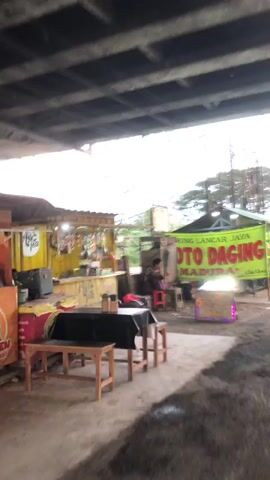 The video depicts a street food stall in Kejapanan, Indonesia. The scene is set under an elevated structure, possibly an overpass, casting shadows and creating a somewhat dim atmosphere. The time of day appears to be overcast, or possibly evening, given the lighting.

The main subject is a person, likely a vendor, sitting at a small stool in front of a food stall. They are wearing a dark brown or maroon top and purple pants. The vendor is engaged in an activity at the stall, possibly preparing food or serving customers, though their specific action is unclear.

Various objects are visible, including the food stall itself, which appears to be a yellow structure with some items on display. There are also chairs and tables for customers, including a red-topped table in the foreground. A green banner with prominent red lettering is displayed behind the vendor, indicating the name of the establishment. Adjacent to this banner, there is a white sign with text. To the right of the vendor, a small stall with purple neon lights displaying "SOTO MADURA" is also visible, suggesting that soto is a specialty. Other items like plastic bags and barrels are scattered around the area.

The overall scene suggests a humble, local eatery. The ground is dirt and uneven, typical of outdoor markets or roadside vendors. The presence of an overpass and the general clutter imply a busy, perhaps less regulated, urban or semi-urban environment. There are no clear indications of the weather, other than the diffused light. The mood is neutral, focusing on the everyday operation of a street food business.

Visible text includes:
- "URUNG LANCAR JAYA" (partially visible on the green banner)
- "SOTO DAG" (partially visible on the green banner, likely part of "SOTO DAGING")
- "MADURA" (on the green banner)
- "SOTO MADURA" (on the stall with neon lights)
- Text on the white sign behind the green banner is too small to read clearly but appears to be official notices or rules.