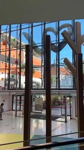 The video captures an indoor scene with large glass windows reflecting the outside. Inside, a wooden tree-like structure with branches is prominently displayed, surrounded by several large wooden posts. In the background, through the windows, a street scene is visible with buildings, palm trees, and colorful circular decorations hanging from the ceiling. Several people are walking around the indoor space. One person in the foreground on the left is wearing a white top and shorts. Further in the background, two individuals are walking side-by-side, with one wearing a light green top and white pants. Another person is walking towards the right. The floor is tiled with a colorful geometric pattern. A pink ribbon or tape is visible across some doorways.