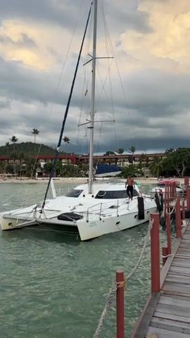 A person stands on a white catamaran docked at a wooden pier. The catamaran has two hulls, a tall mast, and sails furled. The person, wearing dark shorts and no shirt, is looking down at the boat. Another person stands further down the pier, taking a photo with a phone. The water is green and slightly choppy. In the background, a beach is visible with palm trees and buildings. The sky is cloudy with streaks of orange and yellow from the setting sun.