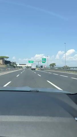 The video is filmed from inside a car, looking forward through the windshield. The car is on a multi-lane highway with clear blue skies and scattered clouds overhead. A green highway sign indicating "FIRENZE" with a downward arrow is visible. A white truck with a logo on its side is in the right lane, further ahead. Other vehicles, including a white bus, are also on the highway. The dashboard of the car is visible in the foreground. The overall scene suggests travel on a sunny day on an Italian highway.