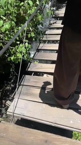 A person wearing brown pants walks across a wooden suspension bridge over a stream. The bridge has wooden planks for a walking surface and metal cables for support. Lush green foliage and trees are visible on either side of the bridge and surrounding the stream. The water in the stream appears to be flowing rapidly, with some small waterfalls visible. The sun is shining, casting shadows on the bridge. Another person is visible in the distance, also on the bridge.
