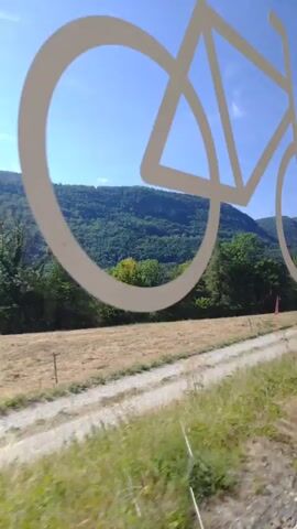 The video shows a view from a moving vehicle, likely a train or car, looking out a window. A white outline of a bicycle is visible in the foreground, obscuring part of the view. The background consists of a landscape with rolling green hills covered in trees, a dirt road, and a clear blue sky. There are areas of dry, yellowed grass and some green foliage. The camera appears to be panning or moving horizontally, giving a sense of travel through the scenery.