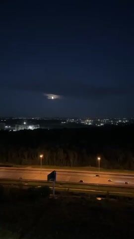 The video shows a nighttime aerial view of a highway and a distant cityscape. The moon is visible in the dark blue sky, partially obscured by clouds. Streetlights illuminate the highway, where a few cars are driving. A dark forest is located between the highway and the city lights. A signpost is also visible in the foreground.
