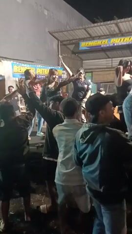 A group of people are gathered in front of a stage. Several women are performing on the stage, some are singing and others are dancing. The audience members are watching the performance and some are dancing as well. The environment appears to be outdoors, possibly at a street event or market, with a building in the background that has banners on it. The lighting suggests it is nighttime.