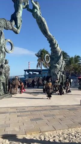 The video shows a group of performers in Playa del Carmen, Mexico dancing in traditional costumes in front of a large sculpture. Most of the performers are men who are shirtless, wearing ornate headwear, and black skirts or coverings from the waist down. The group is accompanied by musicians playing drums. They are dancing on a brick plaza near the beach, a large sculpture, and a small building. The sky is blue and there are some palm trees in the background. A few onlookers are visible sitting on benches.