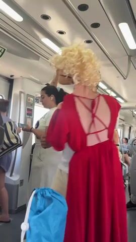 The video shows the interior of a train carriage filled with people. The camera is positioned behind a person wearing a bright red dress with an open back and a blonde curly wig. This person is facing away from the camera, towards the front of the train. Other passengers are visible in the background. To the left, a woman in a light-colored dress is holding a phone. Further left, another woman is visible in a blue top and shorts. To the right of the person in the red dress, a man is standing close to them, partially obscuring their face. The train carriage has fluorescent lighting along the ceiling and handrails. A blue bag is in the foreground, partially obscuring the bottom of the frame. The overall atmosphere appears to be casual, with passengers standing and interacting.