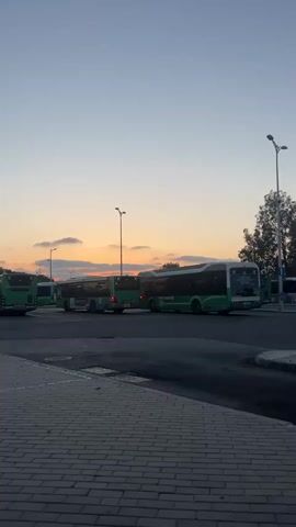 The video captures a serene bus depot or parking area at dusk, under a sky painted with warm orange, pink, and light blue hues near the horizon. Scattered clouds drift above, reflecting the fading daylight.

Several public transport buses, primarily green with white roofs and distinct "X" logos, are visible. One of these buses is slowly maneuvering, its red brake lights illuminating as it turns or backs up. On its rear display, the number "0" is visible, likely indicating a route or status. The bus also features a Hebrew license plate and advertisements in Hebrew along its side. Other similar green and white buses are parked stationary around the depot, also bearing the "X" branding and Hebrew text.

Tall streetlights dot the area, though they are not fully illuminated against the twilight sky. The ground consists of asphalt paving where the buses are parked, and a brick-paved sidewalk in the foreground. Trees are visible in the background to the right. The scene conveys a peaceful atmosphere at the end of the day, consistent with a bus terminal in Kiryat Motzkin, Israel, given the Hebrew text and bus designs.