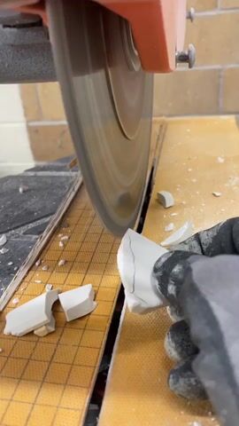 A close-up shot captures a person wearing a gray glove cutting a white, irregularly shaped object with a circular saw. The saw blade, which is spinning rapidly, appears to be making contact with the object. Several small white fragments are scattered around the cutting area, suggesting the object is brittle. The object itself has visible cracks on its surface. The saw is attached to an orange housing unit, and the background shows a tiled surface and a brick wall.