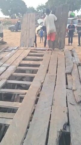The video shows a precarious-looking wooden bridge with large gaps between the planks, revealing the supporting structure beneath.  Several people are on or near the bridge. A man in a light-colored shirt and red shorts stands on the bridge, leaning against a vertical wooden plank, appearing to adjust or examine it. Other individuals are walking around the area, some near vehicles in the background, including a truck loaded with what appears to be sugarcane. The environment seems to be a sandy or dirt-covered area, possibly near a river or other body of water. There are a few trees in the background. The lighting suggests it is daytime, but potentially overcast. People are walking about and seem to be engaged in some activity related to the bridge or vehicles.