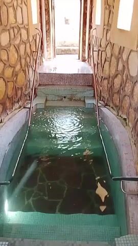 The video shows a narrow indoor pool with steps leading up to a doorway at the far end. The pool is filled with clear water, and the walls are made of stone. Handrails line the sides of the steps. There are small arched windows on the walls. The ceiling is made of wood panels. The doorway at the end opens to a bright outdoor area.