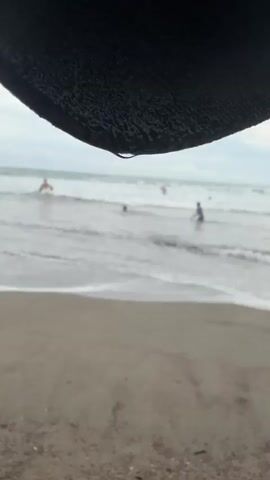 A close-up shot shows a black textured surface with a water droplet hanging from its edge. Below, a blurry beach scene unfolds. Sand occupies the foreground, transitioning to the ocean in the midground. Gentle waves roll towards the shore, and several people are visible in the water, appearing to swim or wade. The sky above is overcast.