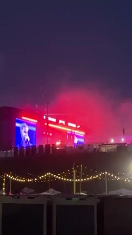 The video captures an outdoor night scene at what appears to be a concert or festival. A stage in the distance is illuminated by dynamic lighting and smoke effects. Large screens on the stage display purple silhouettes, which are visible against alternating green and pink/purple lighting. Dense smoke or fog, colored by the stage lights, billows around the stage area. In the foreground, multiple strands of warm white string lights are strung horizontally. Two separate cube-like structures, each equipped with screens showing similar purple figures, are also illuminated by the changing stage lights and surrounded by colored smoke. A blue flag with a white emblem is visible to the right of one of these cube structures.