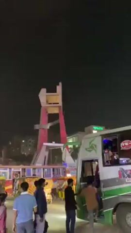 The video shows a busy street scene at night, with a large pedestrian bridge structure illuminated with pink and white lights in the background. Several buses or vans are parked on the right side of the frame, with people standing near them and some appearing to board one. On the left, there are several brightly decorated vehicles, possibly auto-rickshaws. People are walking around, some in groups, and there are illuminated streetlights above. The sky is dark. Overall, the scene captures a lively urban setting in the evening.