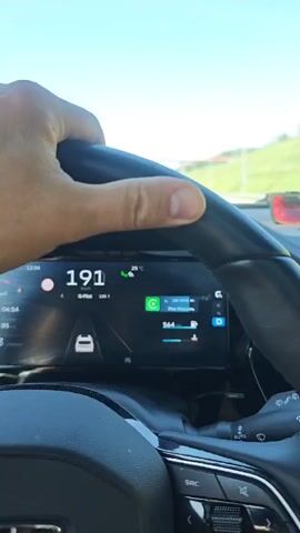 A first-person perspective video shot from inside a car shows the dashboard and part of the steering wheel. A hand is visible gripping the steering wheel. The digital speedometer shows the car accelerating, reaching a speed of 199 kilometers per hour. The digital display also shows the car's temperature as 25 degrees Celsius, the car's fuel level, and information about the car's driver assist system. The road ahead is visible through the windshield. A red car is visible in the right lane ahead. The sky is blue, and green hills are visible in the distance.