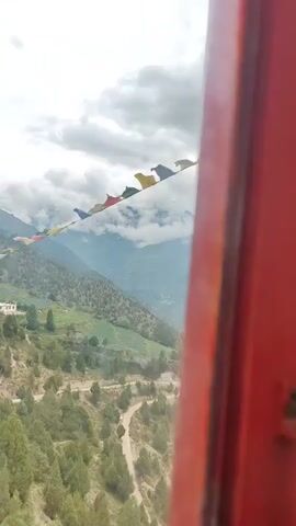 A vertical video begins with a view of a mountainous landscape. Prayer flags are strung across the frame, leading the eye towards a cluster of buildings nestled on a mountainside, with a winding road visible below. The camera pans to the right, revealing more of the rugged terrain and sparse vegetation. The sky is overcast with dramatic clouds that partially obscure the mountain peaks. The video then shifts to a close-up view of a rough, light-colored wall and a section of a dark red, glossy surface, possibly a doorframe. The perspective changes again to reveal a wider vista of the valley, with a river snaking through it and small villages scattered across the slopes. The final shot shows a person's hand briefly entering the frame from the right.