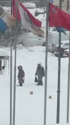 The video shows a snowy outdoor scene with at least two people walking through the snow. The foreground contains a row of flagpoles with various flags, including red and white ones. Snow is falling heavily, and the ground is covered in a thick layer of snow. The people are wearing winter clothing and appear to be moving slowly through the snow. In the background, there are cars covered in snow, suggesting a significant snowfall. The overall environment indicates a wintery, snowy day with limited visibility.