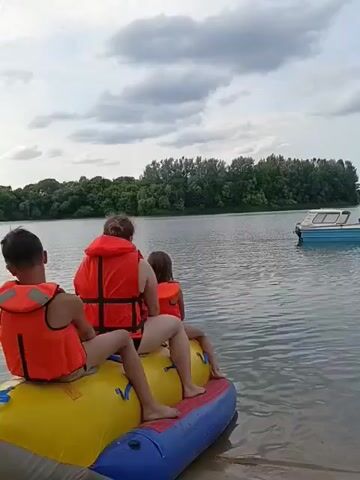 The video begins with a wide shot of three people, two children and one adult, sitting on a yellow and blue inflatable raft at the edge of a body of water. They are all wearing orange life vests. The scene appears to be a calm lake or river on a cloudy day. Trees line the opposite bank in the distance, and a small white boat is visible further out on the water. The raft is partially submerged in the water, and the sand from the shore is visible beneath it. The camera pans slightly to the left, showing more of the water and the surrounding environment. The people on the raft are looking out at the water, and their body language suggests they are relaxed and anticipating an activity. The video ends with a brief shot of the water.
