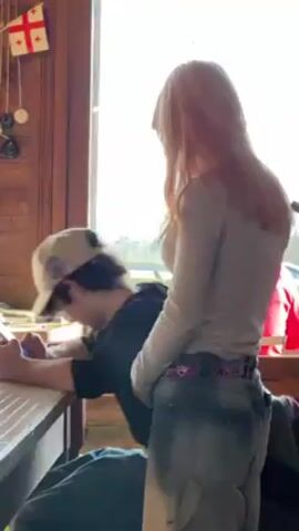 Two people are in a room, likely a cafe or restaurant, with a Georgian flag hanging on the wall. A young man in a baseball cap is seated at a wooden table, looking at his phone. A young woman with long blonde hair stands next to him, looking towards the window. She is wearing a light-colored long-sleeved shirt and faded blue jeans with a colorful belt. The room is well-lit by sunlight coming through the window.