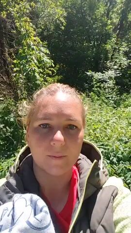 A woman is taking a selfie video in a park. She is wearing a jacket and a red shirt. She is standing on a cracked paved path with trees and foliage in the background. The sky is visible through the trees. The woman has blonde hair pulled back.