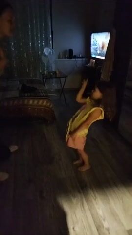 The video shows two people, one adult and one child, standing in a dimly lit room. The adult, on the left, is wearing a dark t-shirt and dark pants. The child, on the right, is wearing a yellow top and pink shorts. The child is facing the adult and appears to be dancing or mimicking the adult's movements. The adult is also gesturing with their hands. In the background, there is a bed with a patterned spread, a fan, and a television screen that is on. The floor is made of dark wood.