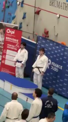 Two young men in judo uniforms are on a podium. One man, with a blue belt, is on the second place step, and the other, with a black belt, is on the first place step. They are facing forward. In front of the podium are several people. Behind the judo players is a blue banner that says “France Judo.”  To the side of the podium is a sign that reads “Orpi - Bienvenue.” Behind the sign is a climbing wall. The sign says “Ville de Brumath” at the top. The floor is blue.