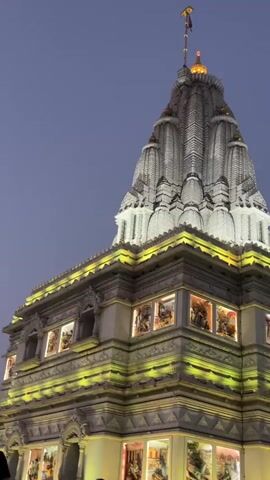 The video shows a Hindu temple illuminated with green lights at dusk. The temple has an intricately carved white tower, and the lower levels are decorated with painted panels. The sky is overcast. A person walks by at the bottom left of the frame, partially obscuring the view.